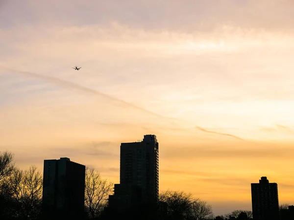 A commercial jet plane flies over residential condominium high rise buildings and trees along Lake Shore Drive and Lake Michigan in Chicago at sunset with beautiful orange cloudy sky beyond.