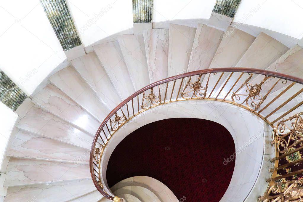 A beautiful fancy marble spiral staircase with red carpet on the bottom and gold colored railing with wood handrails and fluted columns beyond.
