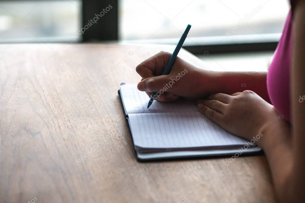 A closeup photograph of mixed race woman wearing a pink shirt and holding a blue pen writing in a modern style note pad sitting at a wood dining table or desk with morning light coming from windows.