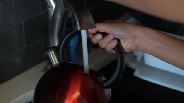 Un primer plano de una mujer afroamericana mientras llena un hervidor de té rojo brillante usado con mango negro redondo y pico de cromo con agua del grifo en el fregadero de la cocina . — Vídeo de stock