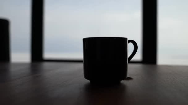 Z bliska z gorącej parze filiżankę herbaty w czarny Kubek ceramiczny siedzący na drewno jadalnia tabeli przed oknami z dużą ilością wpadającego światła białego. — Wideo stockowe