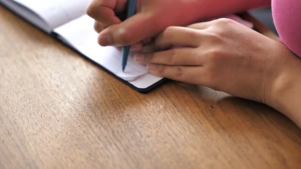 Une vue rapprochée d'une femme noire métissée afro-américaine portant une chemise rose assise à une table à manger en bois et écrivant une note dans son bloc-notes en papier avec un stylo avec ses mains et sa poitrine dans le cadre . — Video