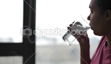 Bir karma yarış Afrikalı-Amerikalı kadın bir pembe tişört soğuk bir bardak buzlu su gelen güneş ışığı ile cam pencere önünde oturan bir yemek masası üzerinden alır.