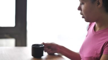 Bir windows gelen güneş ışığı ile ahşap masa karışık yarış Afrikalı-Amerikalı kadın t-shirt bir yemek odası ceviz çay dumanı tüten fincan sıcak alır pembe renkli.