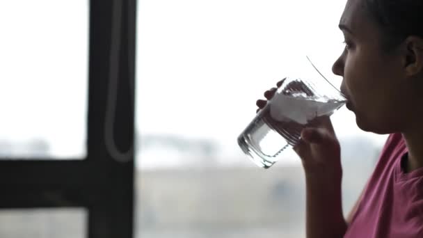 En blandad ras afroamerikanska kvinna i en rosa t-shirt plockar upp ett kallt glas isvatten från ett matbord som sitter framför glasfönster med solljus kommer in. — Stockvideo