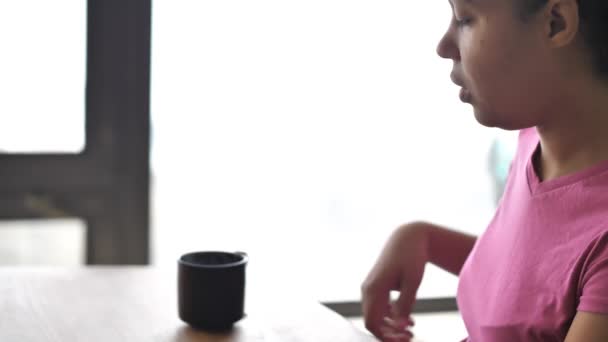 Eine gemischte afrikanisch-amerikanische Frau in rosa T-Shirt nimmt eine heiße, dampfende Tasse Tee von einem Esstisch mit walnussfarbenem Holztisch und Sonnenlicht, das aus einem Fenster dringt. — Stockvideo