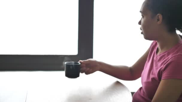 Mieszanej rasy african american Kobieta w różowy t-shirt podnosi gorącej parze filiżankę kawy lub herbaty z orzecha włoskiego jadalni stół z drewna barwić promienie słoneczne padające pod windows poza. — Wideo stockowe
