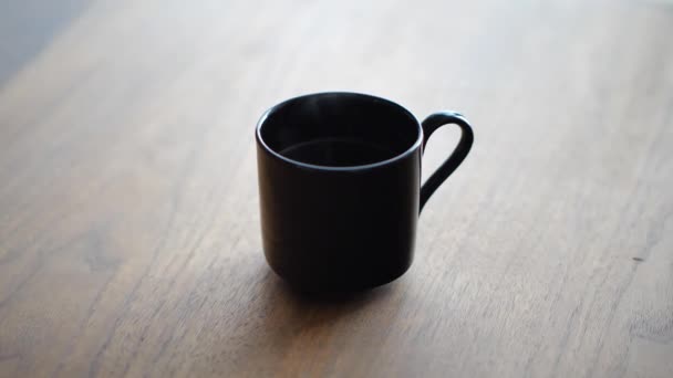 A closeup as a woman's hand picks up a hot steaming cup of tea or coffee from a dining room walnut colored wood table with sunlight coming from a window beyond. — Stock Video
