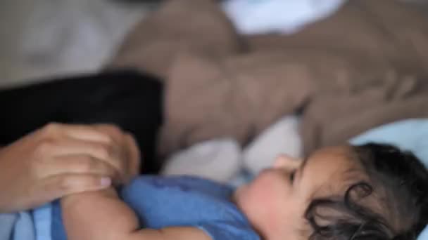 Een gemengd ras Afro-Amerikaanse moeder steekt haar mollige baby op de Wang en kust hem als hij legt op zijn rug in het bed met een pluizige bruine Trooster op de achtergrond. — Stockvideo
