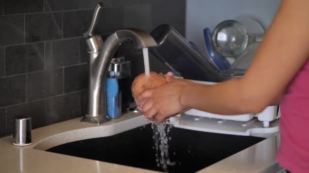 Gros plan d'une jeune Afro-Américaine portant une chemise rose lavant et rinçant une patate douce de couleur orange ou de l'igname dans l'évier de cuisine avec de l'eau du robinet . — Video