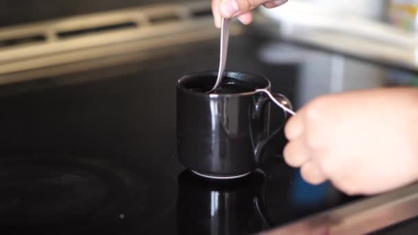 Eine Nahaufnahme der Hände einer afrikanisch-amerikanischen Frau mit gemischter Rasse und einer schwarzen Teetasse, während sie die Teebeutelschnur hält und das dampfend heiße Wasser sanft rührt, während es auf einem Herd steht. — Stockvideo