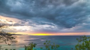 Jamaika'da Ağaçlar tarafından çerçevelenmiş güzel tropikal mavi su ile Atlantik Okyanusu üzerinde gökyüzünde hareket eden mavi, pembe, turuncu ve sarı renkler ile dramatik bulutlar ile muhteşem bir günbatımı timelapse.
