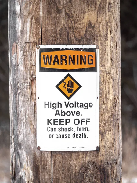 Photograph of a warning high voltage above sign informing people to keep off due to dangers including shock, burn, or death posted on a weathered wood pole or post in a rural area.