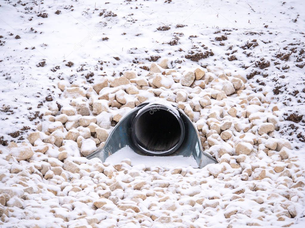 A closeup view of a large storm water drainage culvert pipe emptying into a retention pond set in stone or gravel with snow covering the landscape in rural Wisconsin in the winter season.