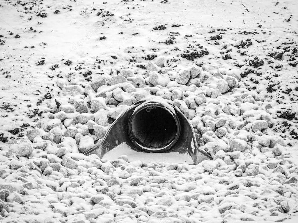 Closeup black and white view of a large storm water drainage culvert pipe emptying into a retention pond set in stone or gravel with snow covering the landscape in rural Wisconsin in the winter season