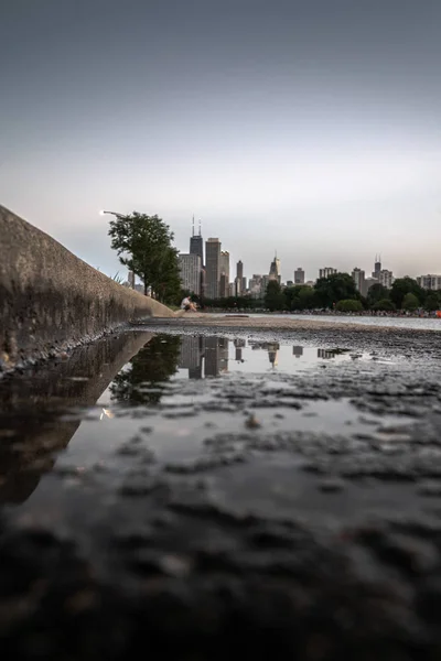 A close up selective focus photograph of a puddle on concrete sidewalk or walkway with curb with the Chicago skyline high rise buildings, a person and tree blurred in the bokeh background beyond.