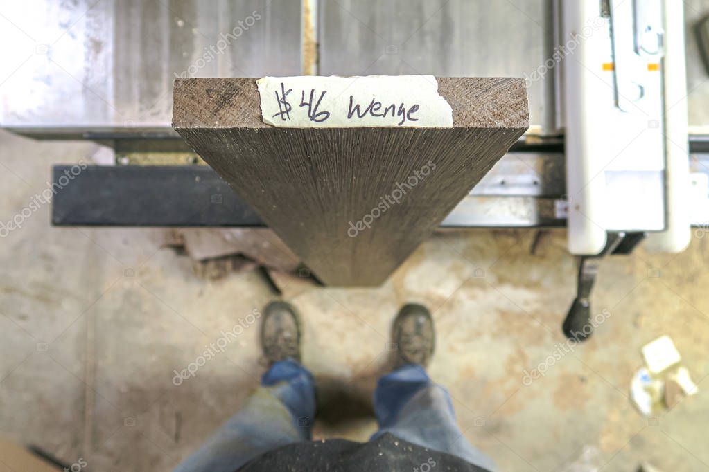 Looking down at a piece of wenge wood with species and cost written on the end of the board with a woodworker's feet in picture below as it leans against a table saw in a woodworking shop.