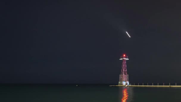 Timelapse de aviones con sus luces encendidas girando hacia el oeste para aterrizar en el aeropuerto mientras reflexionan sobre las tranquilas aguas del lago Michigan mientras la gente camina a lo largo de la costa y el muelle con torre por la noche . — Vídeo de stock