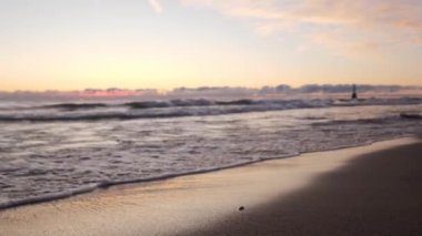 Arkaplanda, kumlu bir sahil şeridine yuvarlanan dalgaların görüntüsü. Renkli, mor ve mavi bir gündoğumu olan gökyüzü ıslak kum ve suyu yansıtıyor..
