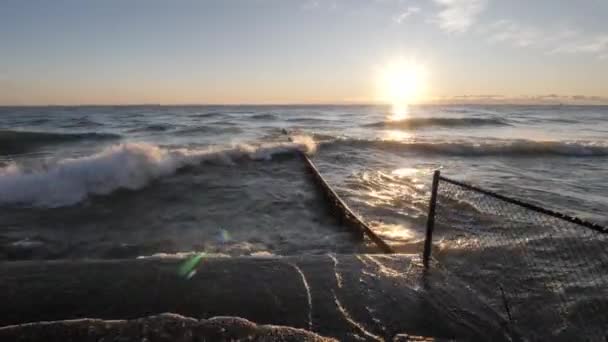 Onde che si infrangono e schizzano sulla costa di cemento e una lamiera d'acciaio che si accumulano nel lago mentre il sole sorge oltre l'orizzonte e si riflette sulla superficie dell'acqua . — Video Stock
