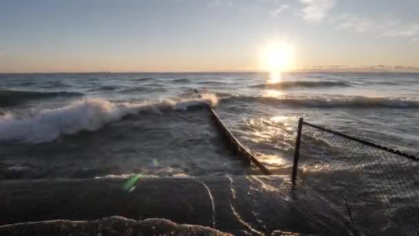 太陽が地平線を越えて上昇し、水の表面に反映しているように、コンクリートの海岸線と湖に伸びる鋼板に衝突し、飛び散る波. — ストック動画