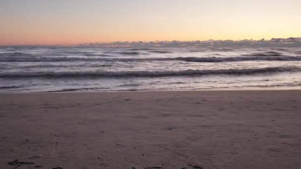 Uma mulher usando sapatos de couro preto e jeans caminha através da moldura deixando pegadas na areia molhada com ondas rolando em uma costa arenosa ao nascer do sol com nuvens rosa e azul no horizonte . — Vídeo de Stock