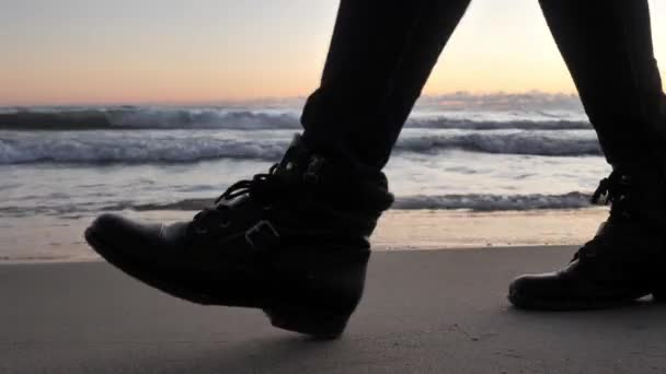 Clip di primo piano delle scarpe alte in pelle nera di una femmina che camminano sulla sabbia lasciando impronte mentre le onde si schiantano sulla costa con nuvole e alba colorata sullo sfondo . — Video Stock