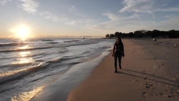 Een gemengde ras vrouw met donker golvend of krullend haar loopt langs de oever van het meer langs de camera op het strand zand als de zon opkomt over het water voorbij als golven rollen in de kust. — Stockvideo