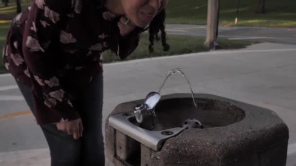 Clip de una mujer afroamericana feliz caminando hacia una fuente pública corriendo y tomando un sorbo del agua fresca y refrescante mientras retiene su cabello . — Vídeo de stock