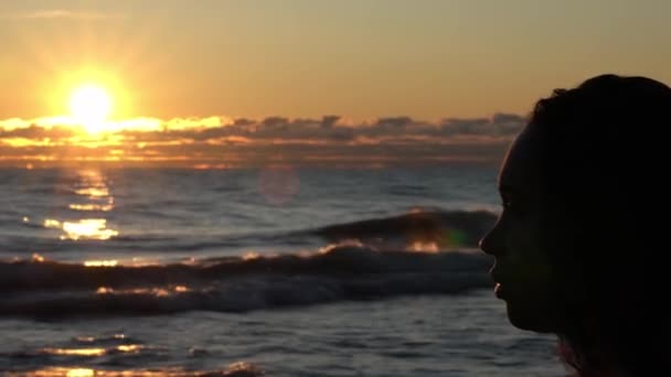 Câmera lenta panning tiro do perfil lateral do rosto de uma mulher bonita em direção ao nascer do sol como o sol reflete cores laranja nas ondas no lago e nuvens logo acima do horizonte . — Vídeo de Stock