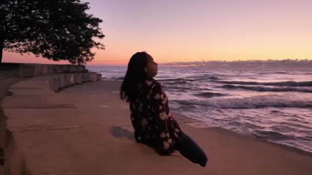 Повільний рух красивої змішаної жінки-гонки з темно-хвилястим волоссям, що тягне себе на кам'яний виступ уздовж озера, коли хвилі врізаються в берегову лінію під час сходу сонця з рожевим небом на горизонті . — стокове відео