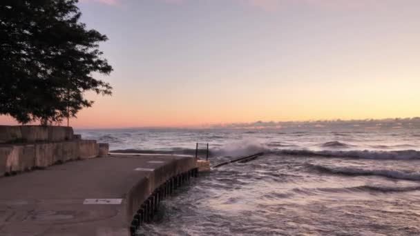 Clip de cámara lenta de las olas del lago Michigan salpicando contra las barreras de acero y hormigón armado cerca de una playa con cielo rosa, azul y naranja al amanecer con nubes en el horizonte y árboles enmarcando la vista — Vídeos de Stock