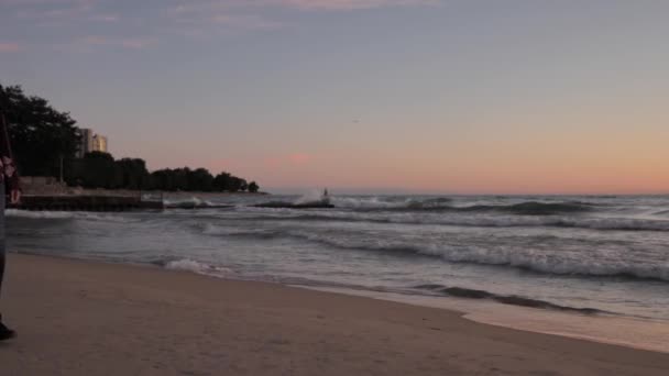 Un clip en cámara lenta de una mujer afroamericana caminando a través del marco en una playa de arena mientras las olas chocan contra la costa del Lago Michigan mientras el sol comienza a levantarse con árboles y edificios más allá . — Vídeos de Stock