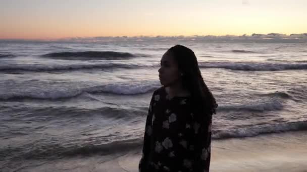 Una vista de cerca de una mujer de raza mixta afroamericana de pie frente al lago Michigan mientras las olas chocan contra la costa al amanecer con el cielo de color naranja más allá mientras mira a su alrededor . — Vídeo de stock