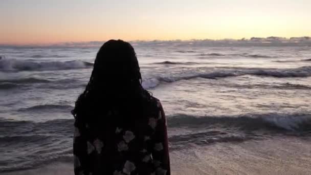 Panning em torno de uma bela mulher mista afro-americana como ondas bater na costa na frente dela como ela está na areia em uma praia ao nascer do sol . — Vídeo de Stock