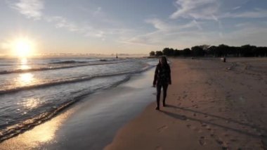 A beautiful African American woman walks along the smooth sand in jeans and long sleeve shirt as the sun on the horizon reflects over the crashing waves along the shoreline Lake Michigan in Chicago.