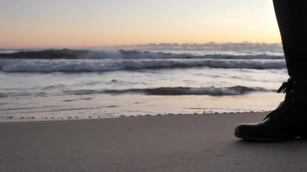 In Zeitlupe gedrehter Close-up-Clip von schwarzen Lederschuhen einer Frau, die über den Sand läuft und Fußspuren hinterlässt, während Wellen mit Wolken und farbenfrohem Sonnenaufgang im Hintergrund in die Küste krachen. — Stockvideo