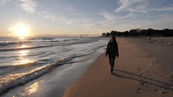 Una hermosa mujer afroamericana camina a lo largo de la arena lisa en jeans y camisa de manga larga mientras el sol en el horizonte se refleja sobre las olas que se estrellan a lo largo de la costa del lago Michigan en Chicago . — Vídeo de stock