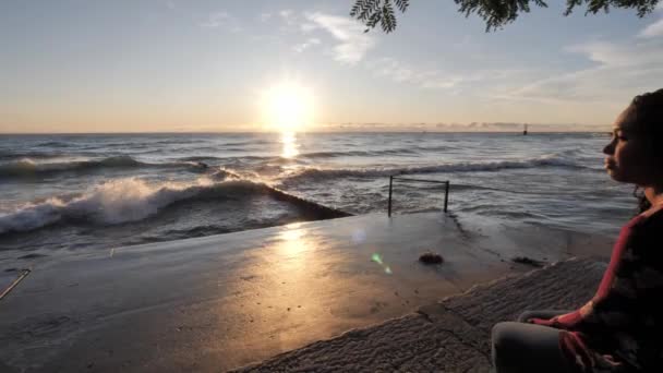 Panning skott från grov vatten och vågor på Lake Michigan vid soluppgången med solen reflekterar på vattnet mot en vacker blandras afroamerikansk kvinna sitter på terrasserade betong strandlinje. — Stockvideo