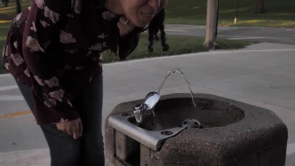 Zpomalený klip šťastné Afroameričanky kráčející k tekoucí veřejné fontáně pití a doušek chladné osvěžující vody, jak si drží vlasy dozadu. — Stock video
