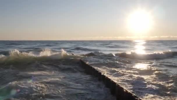 A közelkép lassított felvétel hullámok összeomlik fröccsenő és ütközik egy hullámos acél akadály, ahogy a reggeli nap felkel a horizonton, és tükrözi a víz alatt, és létrehoz egy lencse fáklya — Stock videók