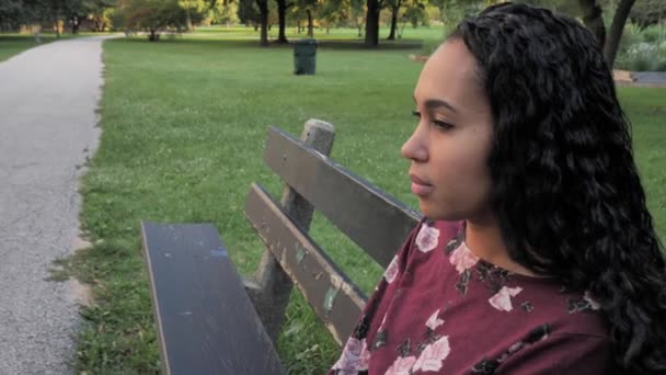 Close-up zijprofiel verticale pan van een mooie Afro-Amerikaanse gemengde ras vrouw zittend op een verweerd hout park bank langs een verhard pad met bomen en vuilnisbak op de achtergrond. — Stockvideo