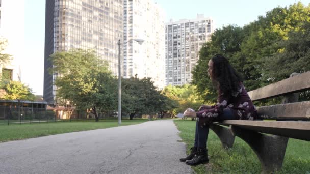 Una joven mujer afroamericana de raza mixta se sienta en un banco de madera y un parque de concreto a lo largo de un sendero mientras cae y recoge un pedazo de papel plegado tirado a ella y lo abre para leerlo mordiéndose el labio. . — Vídeo de stock