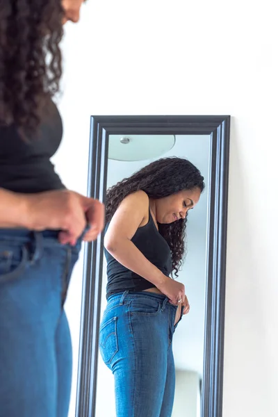 Beautiful African American woman and mother stands in front of a mirror and attempts to try to close her tight fitting blue jeans pants after postpartum diet and weight loss exercise.