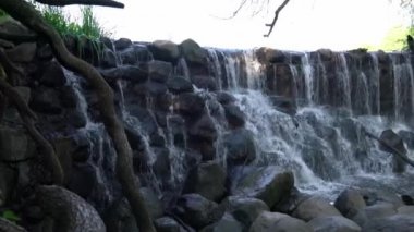 Su tepedeki kayaların ve kayaların üzerine sıçrayıp akarken ve aşağıdaki ağaçlı nehre akarken soldan sağa doğru dönen güzel şelale videosu..