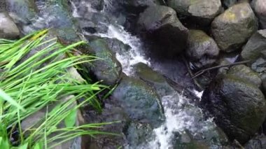 Yakından bakınca, üzerinde çimenler, kayalar ve nehirde kayalar olan küçük bir şelale yapılmış..