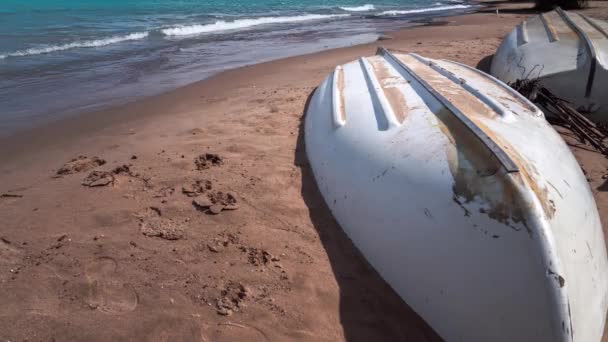 波が砂のビーチに転がる時の経過を閉じます 茶色の砂の中に部分的に埋葬された前景の上に風化した白い列のボートが逆さま — ストック動画