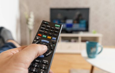 Elinde uzaktan kumandası olan adam akıllı televizyonu izliyor ve uzaktan kumandanın düğmesine basıyor. Uzaktan kumanda yakın planda.