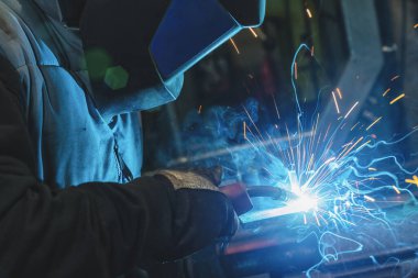Güçlü adam kaynakçıdır, kaynak maskesi ve kaynakçı derisi içindedir. Metal bir ürün, garajda kaynak makinesiyle kaynaklanır. Mavi kıvılcımlar yanlardan uçar. 