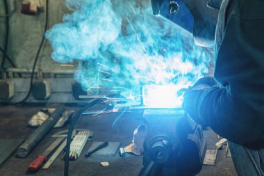 İnşaat eldivenleri ve kaynak maskesiyle kaynakçılık yapan bir kaynakçı atölyede bir kaynak makinesi metali ile çalışıyor.